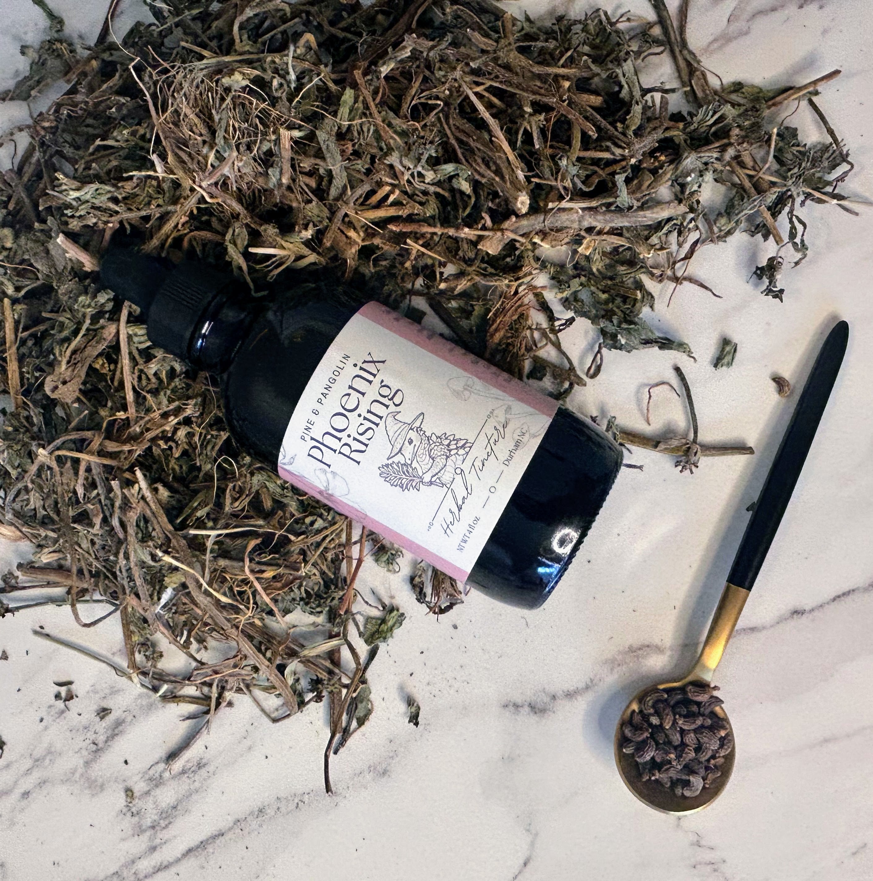 A bottle of Phoenix Rising tincture surrounded by dry herbs and a dropper with a measurement marking, placed on a marble surface. Small batch holistic traditional Chinese medicine and southern folk craft in Durham, NC. 